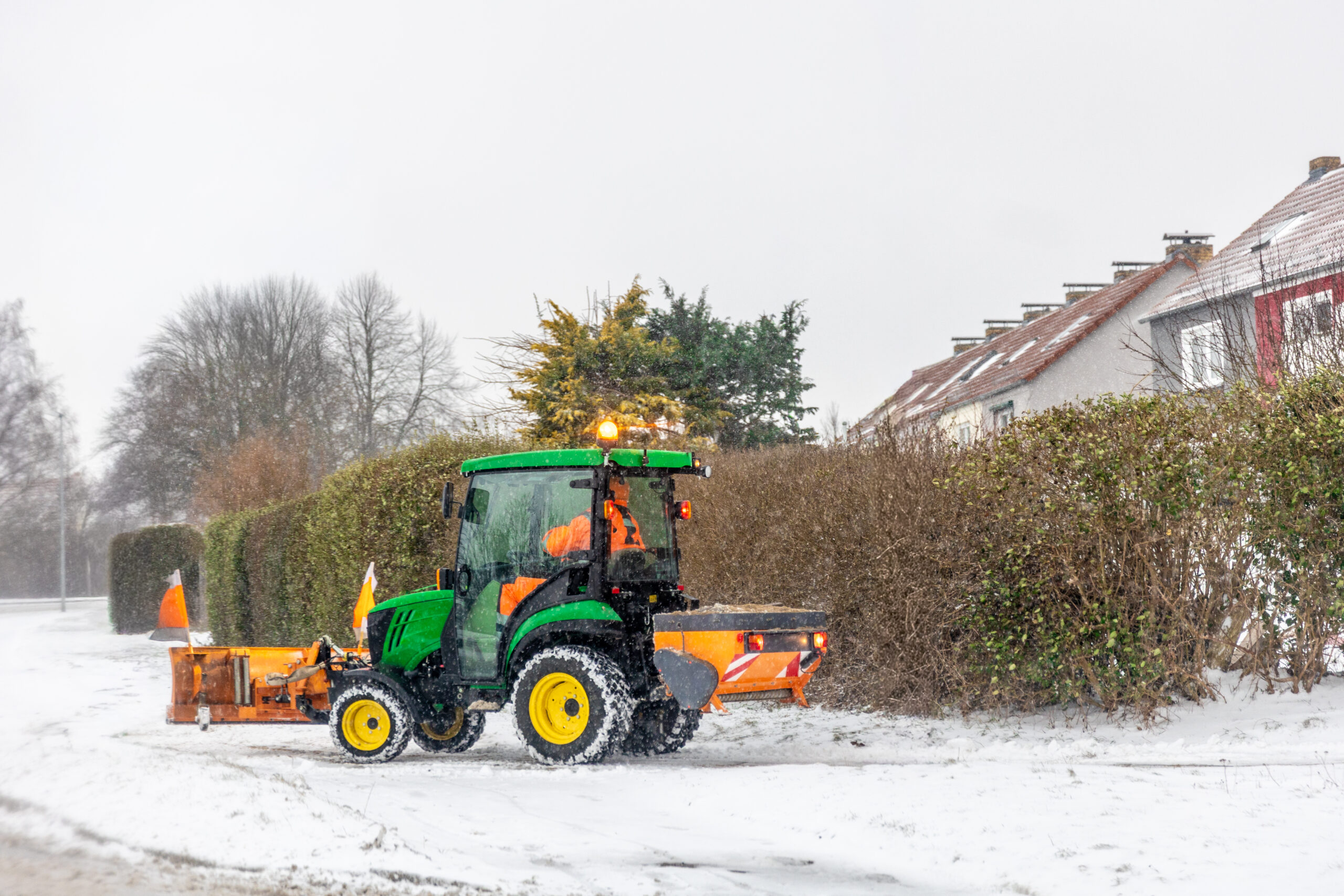 Traktor mit Schneepflug und Streueinrichtung beim Winterdienst auf einer verschneiten Straße.